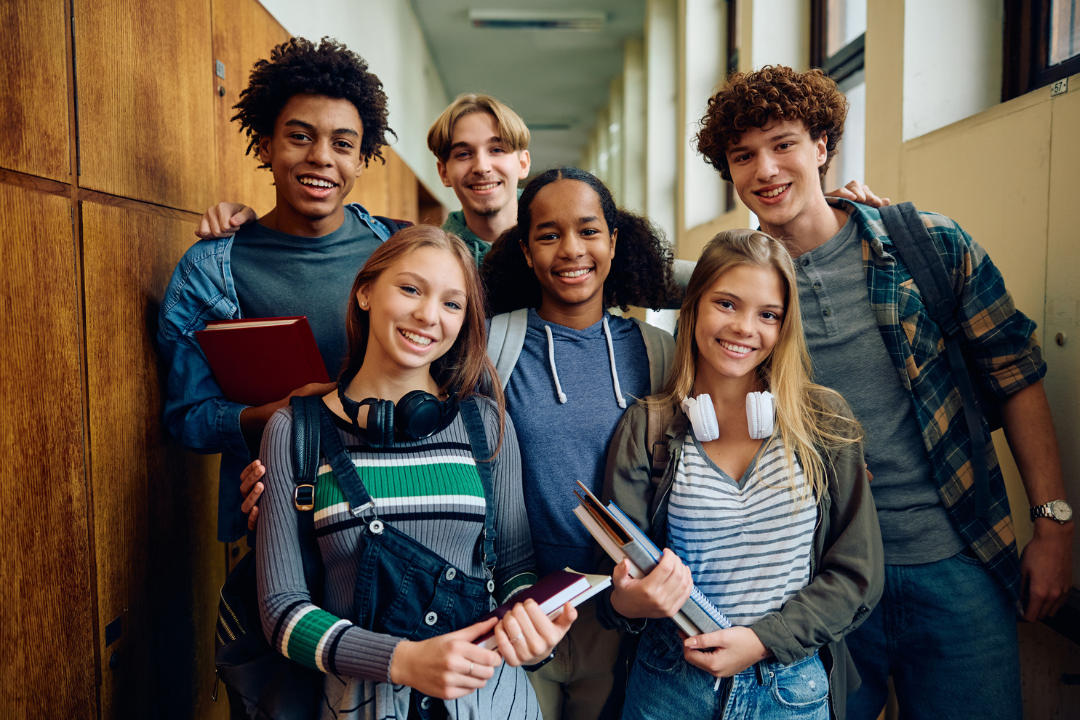 Een groep van 6 jongeren staan lachend in een schoolgang voor kluisjes. Ze houden hun schoolspullen in hun hand.
