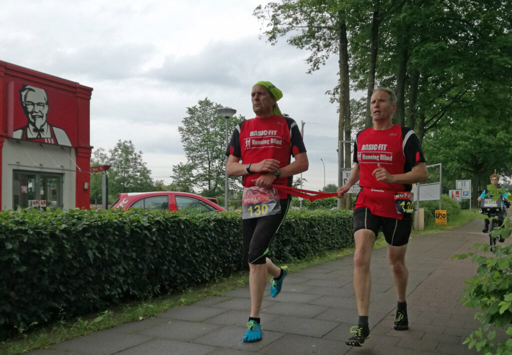John Stoop is aan het hardlopen met een begeleider die hem middels een lint meeneemt. 