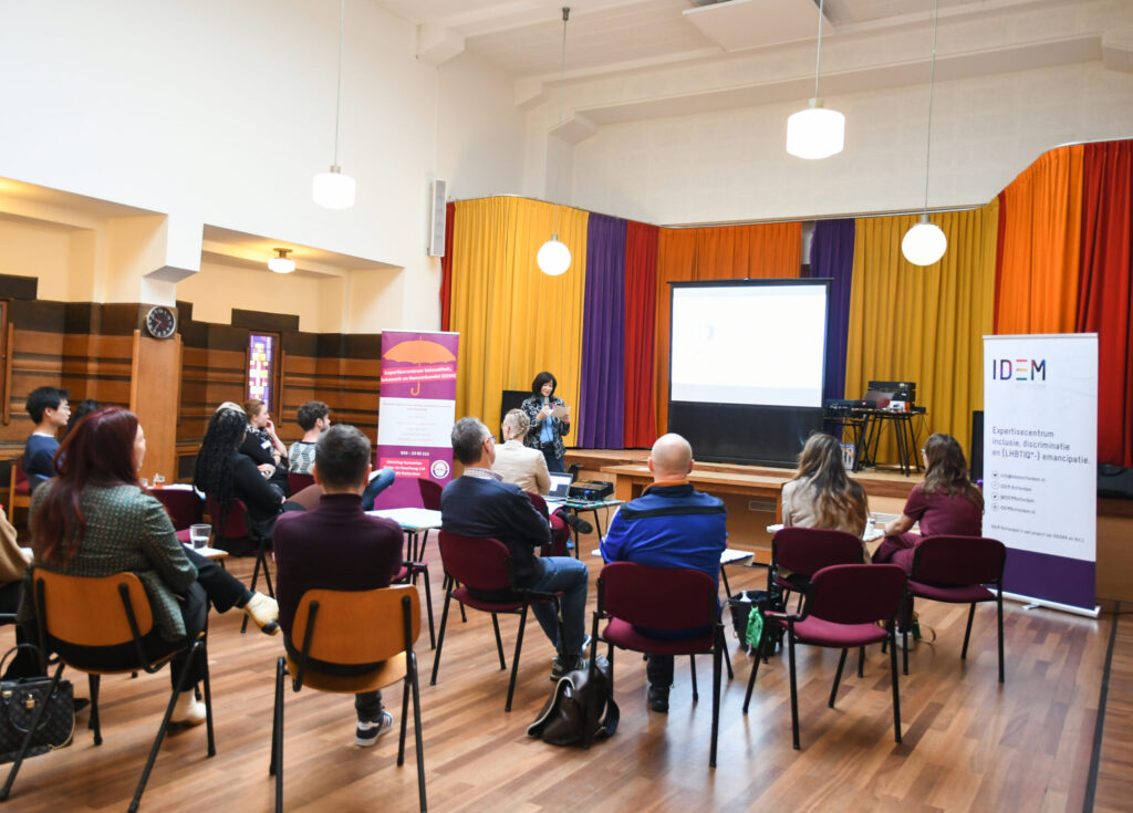 Een zaal vol met mensen die zitten op stoelen, met hun gezicht naar een groot scherm gericht waar de spreker door de microfoon praat.