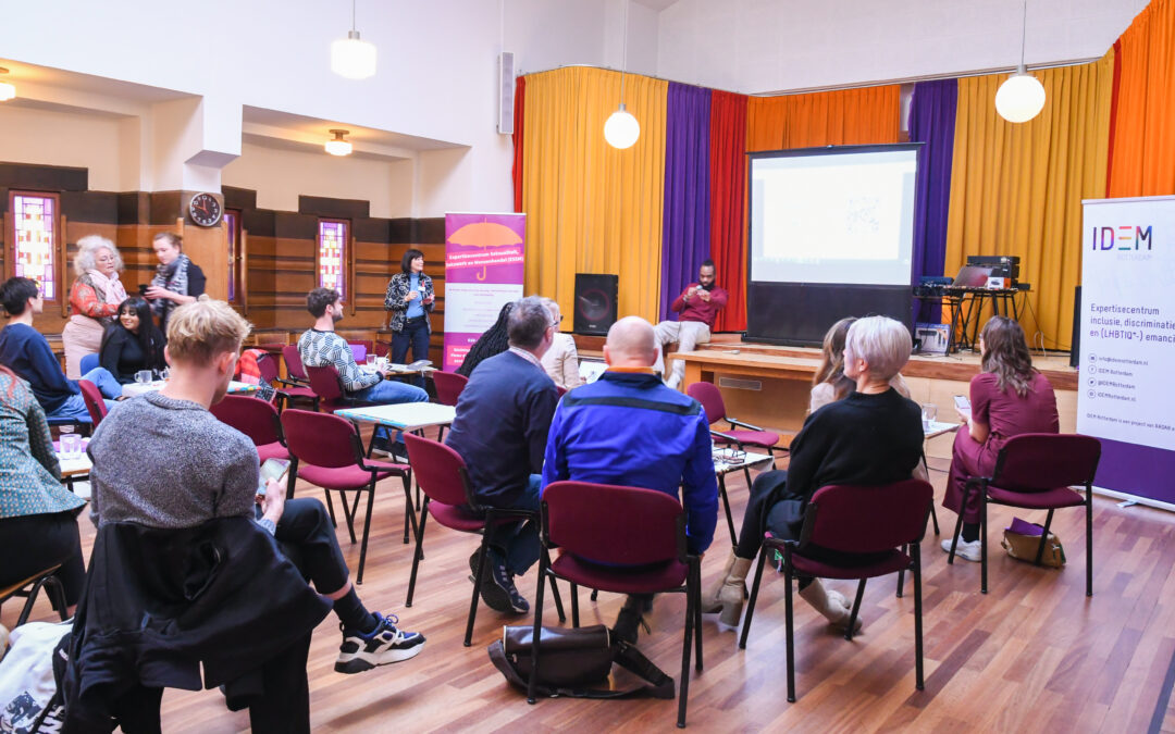 Een zaal waar mensen op stoelen zitten en naar een scherm kijken waar een spreker presenteerd.