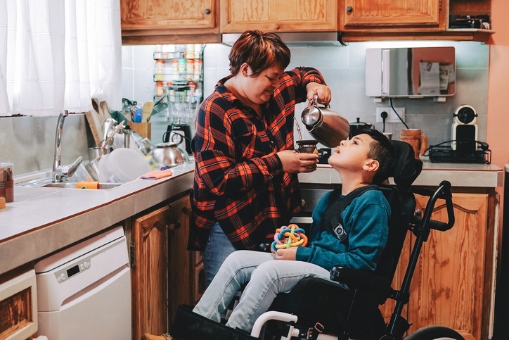 Een ouder zorgt voor haar kind. Het kind zit in een rolstoel. Ze staan samen in een keuken.
