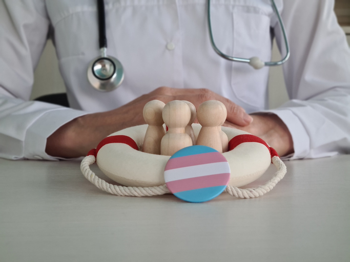 Een arts zit aan zijn bureau. Voor hem zitten houten poppetjes in een reddingsboei. Voor deze poppetjes is een button met de transgender vlag.