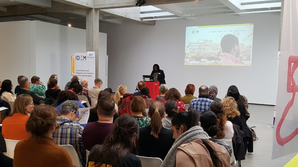 Een groep mensen zit in een zaal op stoelen en kijken naar de presentator. Op de achtergrond wordt een presentatie geprojecteerd op een witte muur.