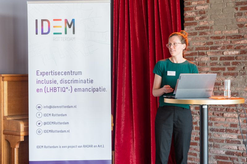 Een spreker staat aan een statafel en spreekt vanachter diens laptop. Naast hen staat een roll up banner van IDEM Rotterdam.