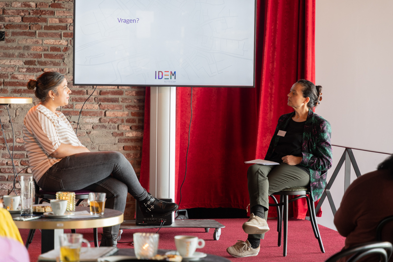 Twee sprekers zitten op stoelen voor het grote tv scherm en zijn in gesprek met elkaar.