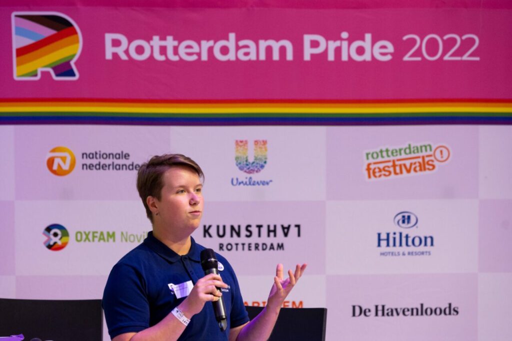 Een spreker spreekt in de microfoon en staat voor een scherm over Rotterdam Pride 2022.