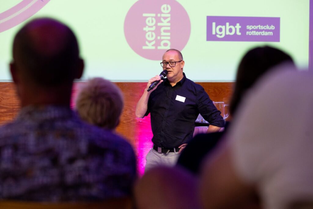 Een spreker staat op het podium en houdt een microfoon in de hand. Het publiek is vanaf de rug te zien.