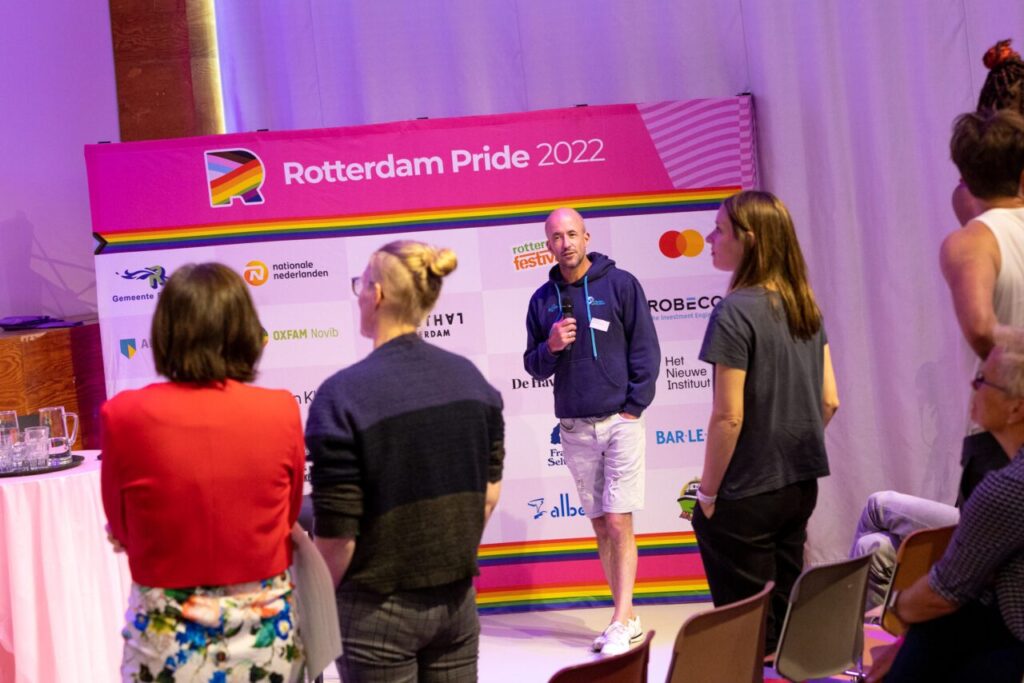 Een spreker is te zien met een microfoon in de hand. Het publiek is te zien vanaf de rug.