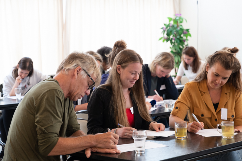 Drie personen zitten aan een tafel en schrijven met pen op papier.