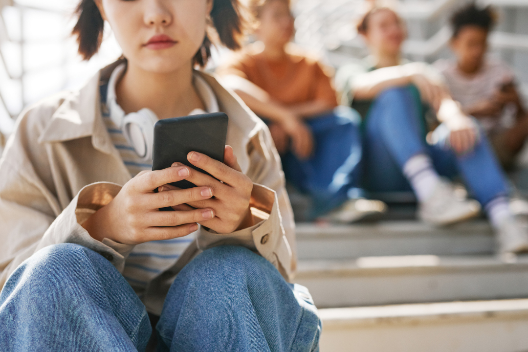 Een persoon zit met de telefoon in de handen op een trap. Op de achtergrond zijn 3 andere personen te zien.