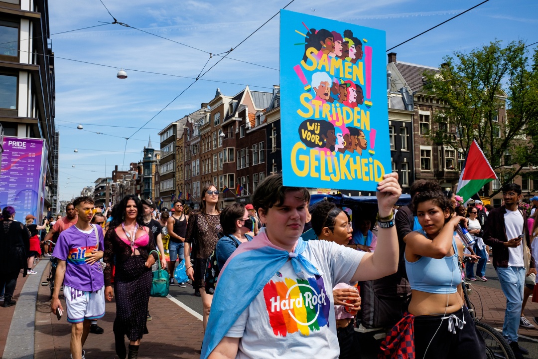 Een stoet mensen loopt door het centrum van Amsterdam tijdens de Women's March 2025. Op de voorgrond houdt iemand een protestbord omhoog dat leest: samen strijden wij voor gelijkheid.