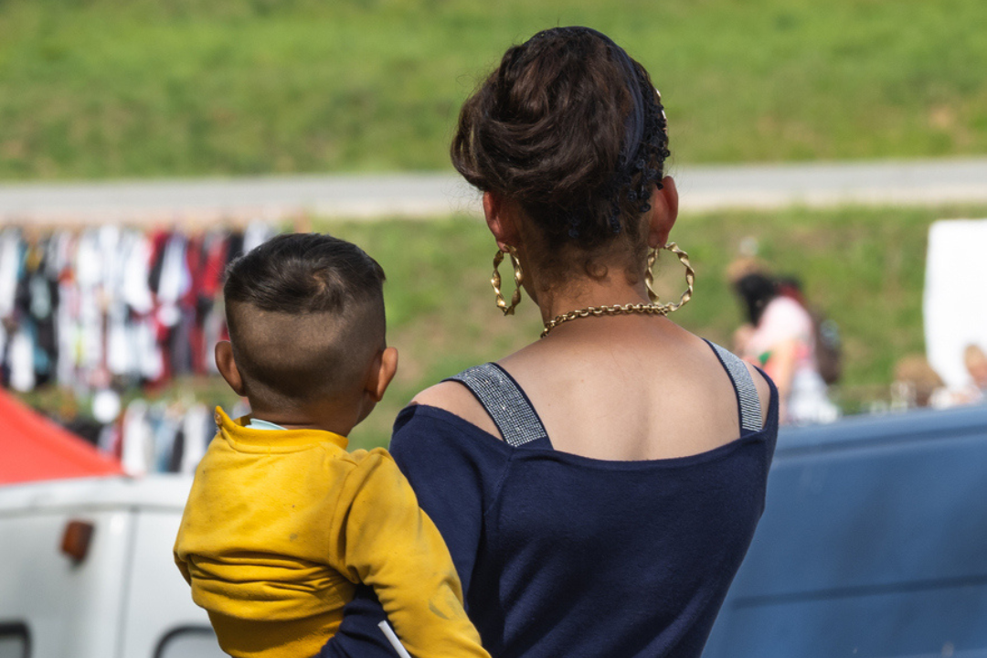 Een Romani vrouw, gezien vanaf de rug, houdt haar zoontje vast op de arm tegen haar heup.