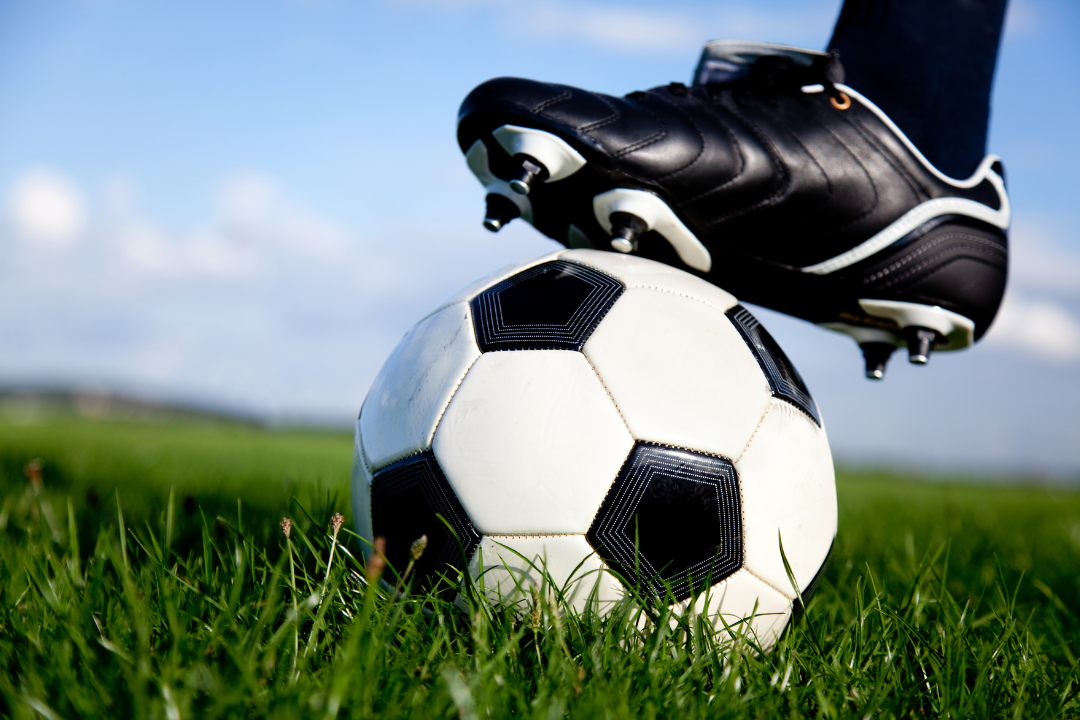 Een voet met voetbalschoen aan staat op een voetbal op een voetbalveld.